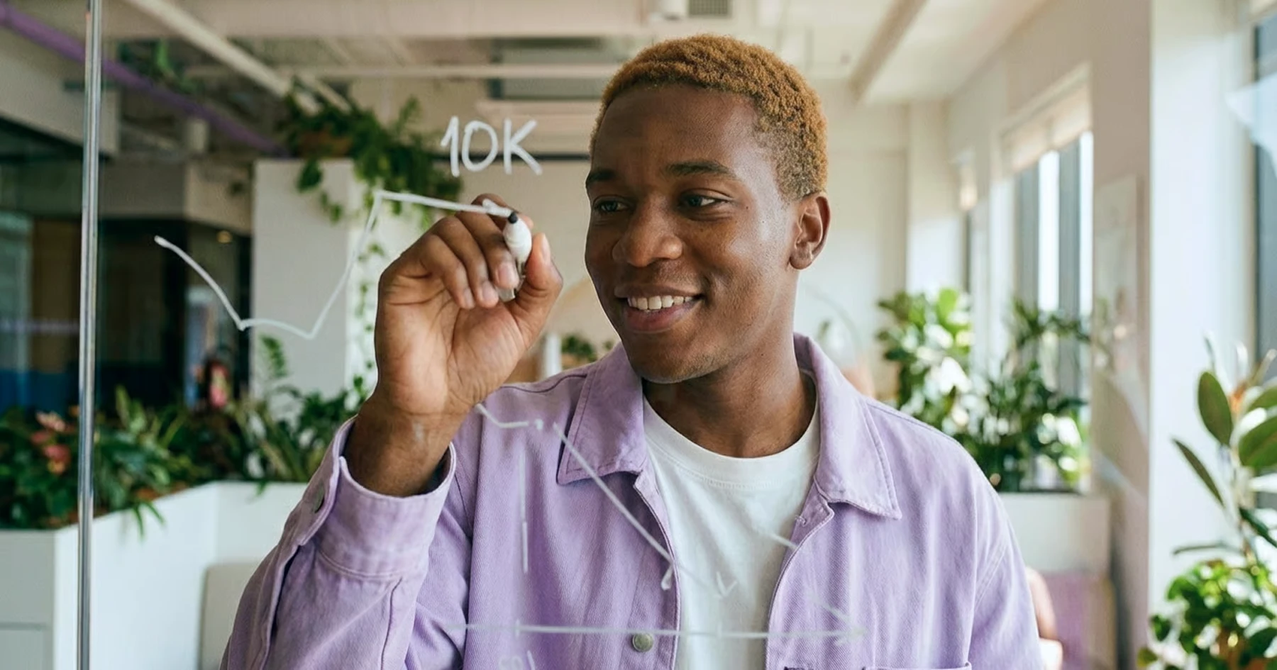 A smiling man in a lilac jacket drawing a rising line and the figure 10K on a glass wall in a bright office full of plants